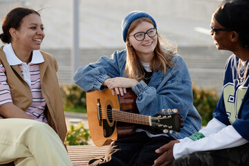 Group of multiethnic teenagers sitting outdoors, smiling and talking while young Caucasian girl with glasses and beanie holding acoustic guitar, enjoying casual conversation together