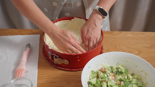 Pressing puff pastry dough into a red baking pan. A woman forms a crust or base for a homemade quiche. The process of making a savory tart shell or pie crust from scratch.