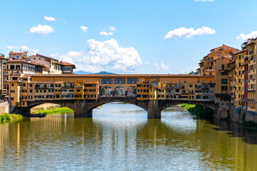 Historic Ponte Vecchio bridge in Florence, Italy, spans the Arno River with charming shop-lined architecture under a clear blue sky on a summer day.