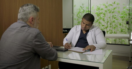 Obraz premium Male doctor writing medical notes during consultation with middle-aged patient seated at desk in clinic, both focused on conversation and paperwork