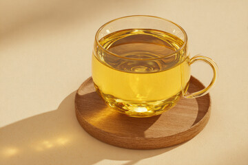 Transparent cup of tea on a wooden coaster, bathed in warm sunlight, casting a soft shadow. Simple elegance.