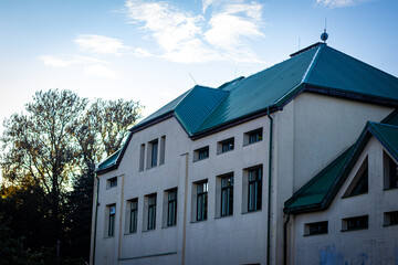 A beige institutional or public building with a green metal roof and numerous rectangular windows, set against a blue sky and surrounded by tall trees.