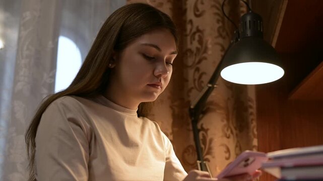 Student girl is happy with a message on her phone. A student is doing homework and is distracted by a pleasant message on her phone that makes her feel happy and joyful
