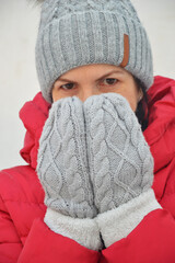 Portrait Caucasian woman in red winter jacket, gray knitted hat with fur pompom, warm knitted mittens on white background. Face covered with hands from cold.