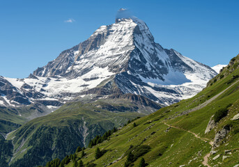 Fototapeta premium Majestic snow-capped mountain peak illuminated by bright sunlight with lush green slopes below under a clear blue sky