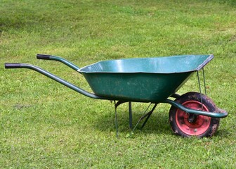 Green wheelbarrow with metal frame and red wheel standing on lawn. Gardening and farming equipment for rural countryside lifestyle.