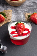 Glass cup with strawberry jelly, cream, and fresh strawberry on top, placed on a dark slate surface with whole strawberries around.