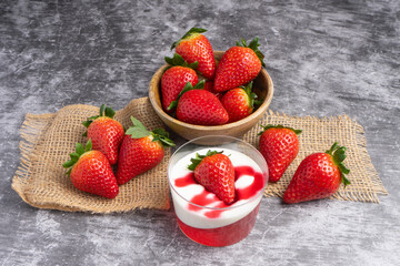 Glass cup with strawberry jelly, cream, and fresh strawberry on top, placed on a dark slate surface with whole strawberries around.