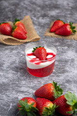 Glass cup with strawberry jelly, cream, and fresh strawberry on top, placed on a dark slate surface with whole strawberries around.