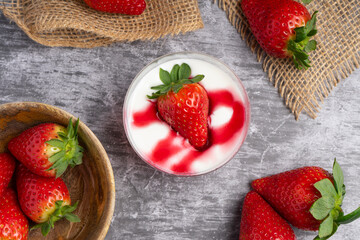 Glass cup with strawberry jelly, cream, and fresh strawberry on top, placed on a dark slate surface with whole strawberries around.