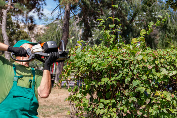 Professional gardener trimming bush with a brush cutter. Garden worker cares of plant cutting bush, mowing grass. Park maintenance