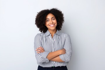 A confident woman with curly hair and a striped blouse stands with her arms crossed, smiling warmly. The setting features a plain backdrop that emphasizes her cheerful demeanor
