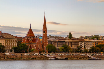 View of Danube riverbank on Buda side featuring Szil&aacute;gyi Dezső Square Reformed Church with red brick neo-Gothic architecture, Zsolnay tile roof, and reflection in pre-sunset sky. Budapest, Hungary.
