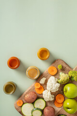 Fototapeta na wymiar Flat lay of fresh vegetables, fruits, and colorful purees in jars on pastel green background with wooden board