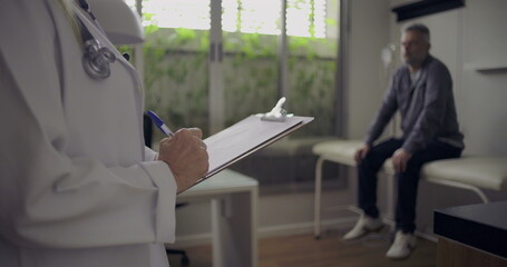 Closeup of female doctor holding clipboard and pen as middle-aged male patient waits in exam room background