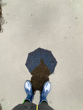 A reflection on a water puddle of a person holding an umbrella during a rainy day and a view of the rain boots for inclement weather concept with copy space. 