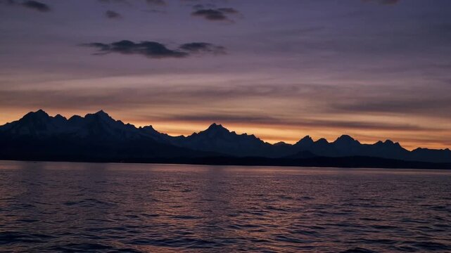 The sun descending behind range of tall and majestic snow-capped peaks across large body of water creating colorful evening sky