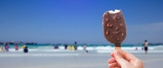 Chocolate covered ice cream bar in the summer at the beach, during the holidays. Hand holds delicious chocolate and nuts almond ice cream bar on a stick.