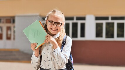 Happy schoolgirl holding notebooks and smiling in front of school building, ready for studying and learning new things