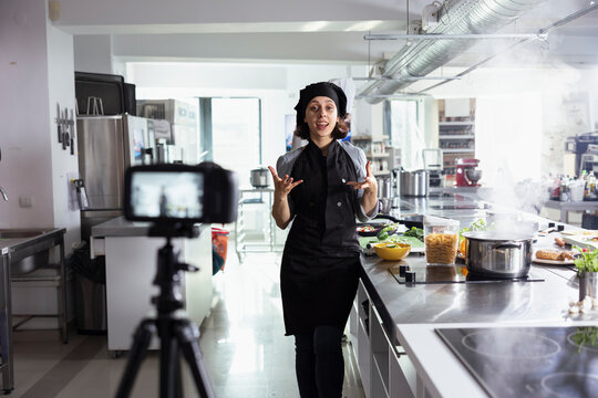 Professional chef in uniform recording a cooking tutorial in a modern kitchen, sharing expert techniques and recipes for an online audience. Woman filming every step of food prep.