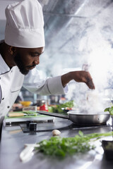 African american head chef sprinkles rosemary over a gourmet dish slow cooked on the stove, balanced gastronomy with fresh herbs. Kitchen counter filled with raw materials and fresh ingredients.