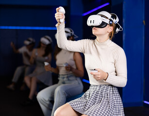 Cheerful emotional young female in VR goggles sitting in virtual reality room with group of friends, manipulating objects or shooting at targets with gaming controllers in hands..