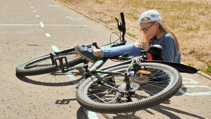 Young female cyclist tending to scraped knee beside fallen bicycle on sunny suburban street pavement