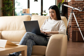 Naklejka premium Focused female entrepreneur sitting on couch with laptop in lap, reviewing business analytics. Asian woman concentrating on digital device, browsing online and updating personal schedule.