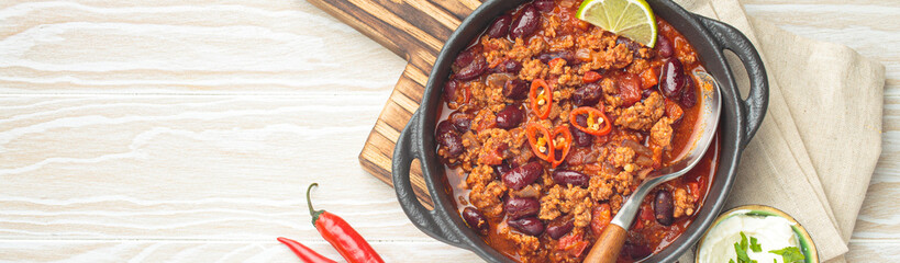 Traditional mexican chili con carne with lime, red chili peppers, and sour cream in a cast iron pan on a wooden board, white background, copy space