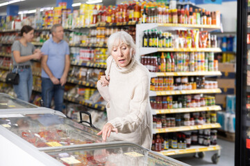 Casual positive aged female buyer standing in front of glass refrigerated display case, looking at assortment of foods arranged inside..
