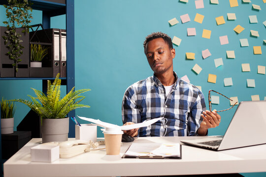 Male consultant sitting with papers in his hand, feeling overwhelmed by amount of business project workload on his desk. Stressed finance advisor holding his glasses and looking at documents.