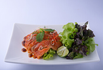 A beautiful orange salmon salad, topped with a dressing, peppermint leaves, chilies, and lime, served with greens on a white plate against a white background.