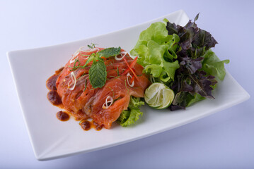 A beautiful orange salmon salad, topped with a dressing, peppermint leaves, chilies, and lime, served with greens on a white plate against a white background.