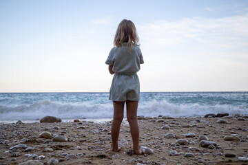 Barefoot blonde woman standing on rocky shoreline, contemplating ocean waves washing against coastal landscape under peaceful sky