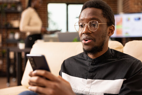 Closeup of black male accountant looking at smartphone with surprised expression, reacting to good news about startup company finances. Shocked businessman with glasses holding his mobile device.