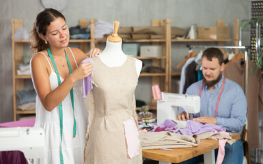 Dedicated young female dressmaker carefully constructing linen dress on mannequin in cozy sewing workshop for custom designs with male teammate working on sewing machine in background..