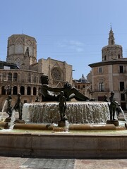 Central square with Fuente del Turia, Turia Fontaine in the historical center of Valencia, Spain. European Gothic Valencia Cathedral against blue sky on a sunny day in Plaza de la Virgen