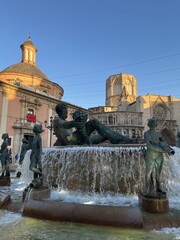 Central square with Fuente del Turia, Turia Fontaine in the historical center of Valencia, Spain. European Gothic Valencia Cathedral against blue sky on a sunny day in Plaza de la Virgen