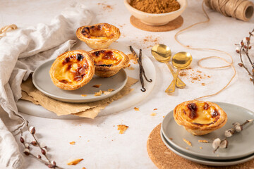 Several Pastel de Nata are arranged on plates with cinnamon, vanilla beans, and small golden spoons.
