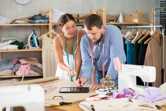 In professional fashion studio, focused young male dressmaker sketching new garment on tablet while inspired female colleague sharing ideas, collaborating on design