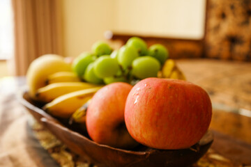 Assorted fresh fruits including grapes, bananas, apples, and pear in a wooden bowl on a decorative table with patterned sofa. 