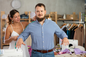 Young seamstress skillfully taking measurements for male client in modest sewing studio, preparing for custom-made clothing order..