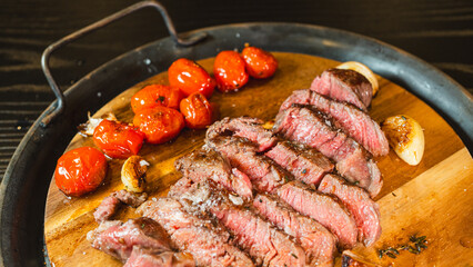 Juicy sliced medium rare ribeye steak served on a wooden board with roasted garlic, fresh thyme, and cherry tomatoes. Ideal for gourmet food content and recipes.
