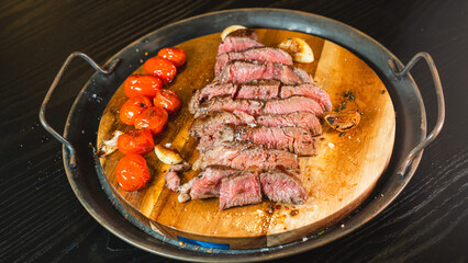 Juicy sliced medium rare ribeye steak served on a wooden board with roasted garlic, fresh thyme, and cherry tomatoes. Ideal for gourmet food content and recipes.