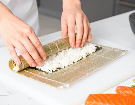 Hands rolling sushi with bamboo mat and rice on seaweed, fresh salmon slices on side ready for preparation in clean kitchen setting