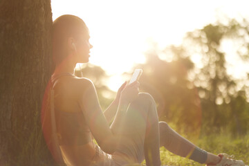 Young woman with headphones relaxing under a tree, enjoying music on a smartphone during sunset, embodying calmness and serenity in nature