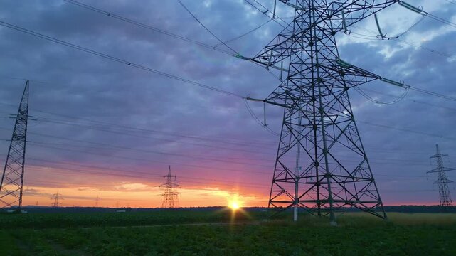 Power lines and pylons silhouetted against a vibrant sunset in a rural field, symbolizing sustainable energy infrastructure and harmony with nature, captured in a long shot.