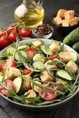 Tasty salad with croutons, cucumbers, tomatoes and arugula on black table, closeup