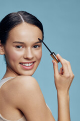Fototapeta premium Young woman applying mascara for a beauty look, showcasing radiant skin and a cheerful expression against a light blue background