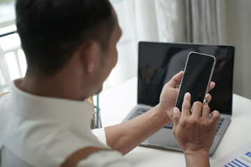 Businessman using smartphone while working on laptop at home office, concept of digital lifestyle, remote work, mobile technology, and modern communication.